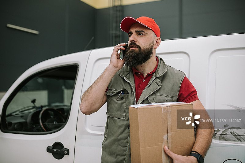邮递员站在他的送货车前，一边打电话一边拿着包裹图片素材