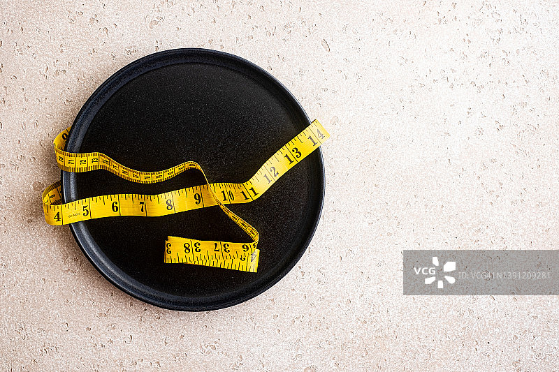 混凝土背景上的空黑盘子和黄色卷尺的俯视图。健康生活方式，节食概念。图片素材