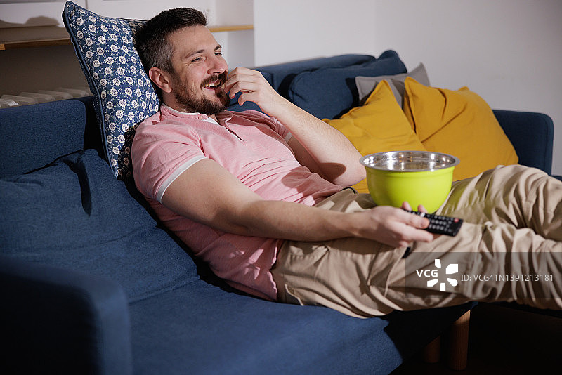 快樂的男人坐在沙發上，抬著腳在家裡吃零食看電影图片素材