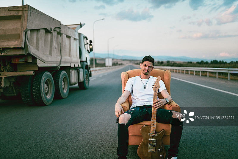 吉他手坐在高速公路的椅子上，卡车经过图片素材