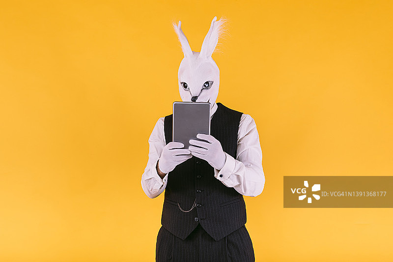 伪装者：穿西装、马甲和领带的兔子面具人在黄色背景下看平板电脑图片素材
