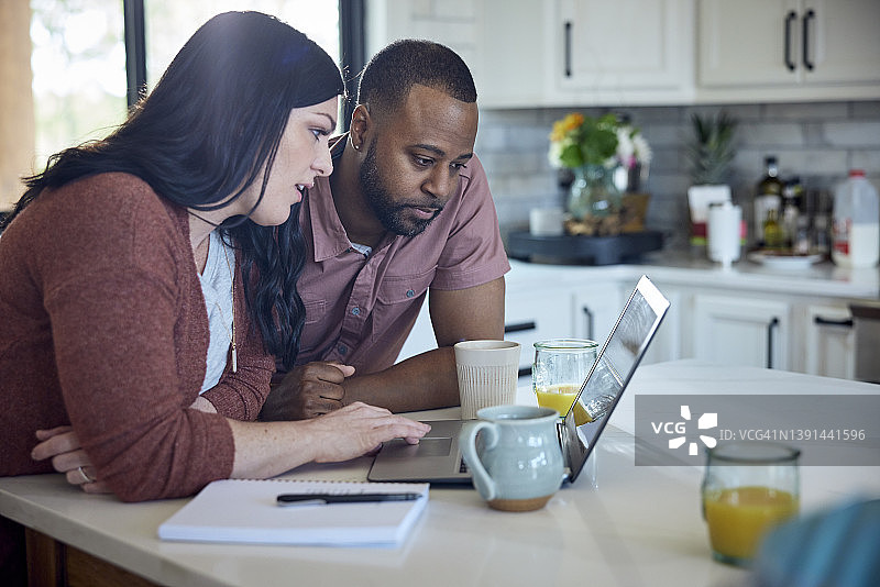 白人女子和非裔男子在厨房的柜台边一边吃早餐一边用笔、纸和笔记本电脑工作图片素材