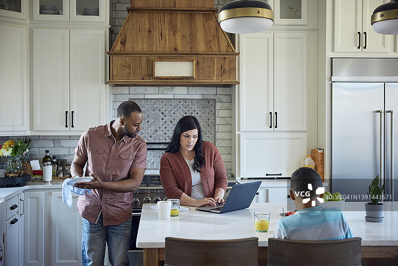 非裔美国父亲洗碗，白人母亲使用笔记本电脑工作，年幼的儿子吃早餐图片素材
