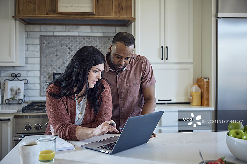 白人女子和非裔男子在厨房的柜台边一边吃早餐一边用笔、纸和笔记本电脑工作图片素材