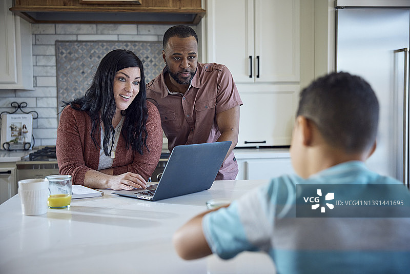 白人妈妈和非裔美国人爸爸与小儿子一起坐在厨房的餐桌旁，用笔、纸和笔记本电脑工作图片素材
