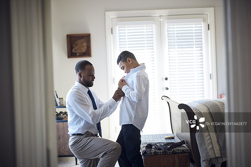 非裔美国父亲帮助混血儿子在卧室里穿衬衫和领带图片素材
