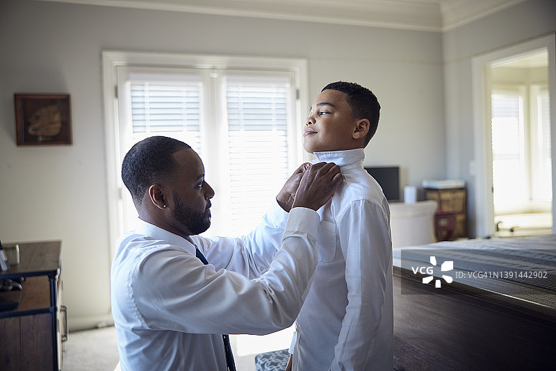 非裔美国父亲帮助混血儿子在卧室里穿衬衫和领带图片素材