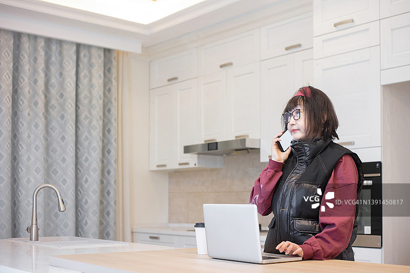 一位东亚女人在厨房里使用笔记本电脑和手机图片素材
