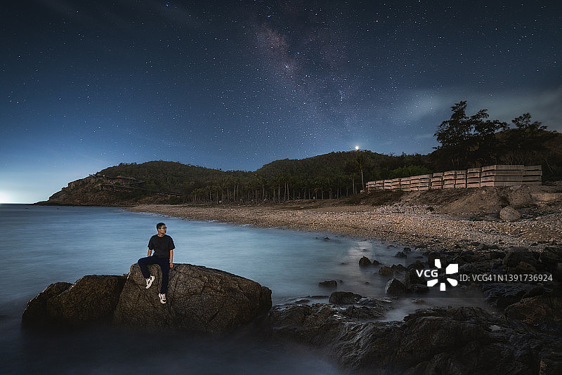 年轻人夜晚坐在海边礁石上看夏季银河图片素材