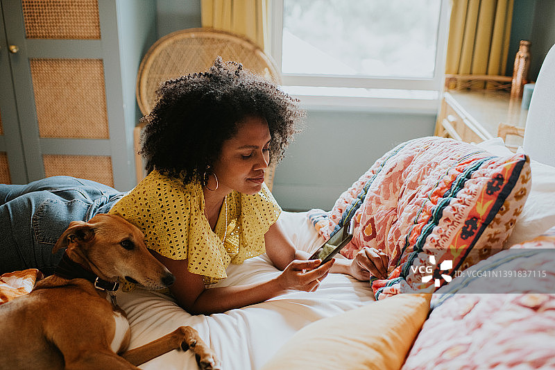 漂亮女人在时尚、色彩鲜艳的卧室里俯卧在床上，她的狗依偎在她旁边，她正在使用手机图片素材