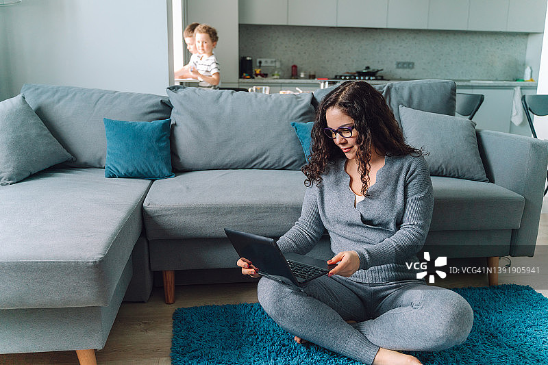 中年女商人在家里的地板上用笔记本电脑工作，背景是两个孩子图片素材