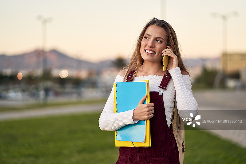 长发女学生提着文件夹和笔记本，背着包，在日落时分公园的草地上用智能手机交谈图片素材