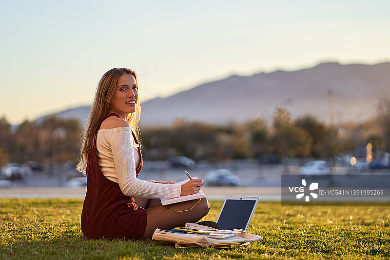 微笑的女人在公园学习时在笔记本上写作图片素材