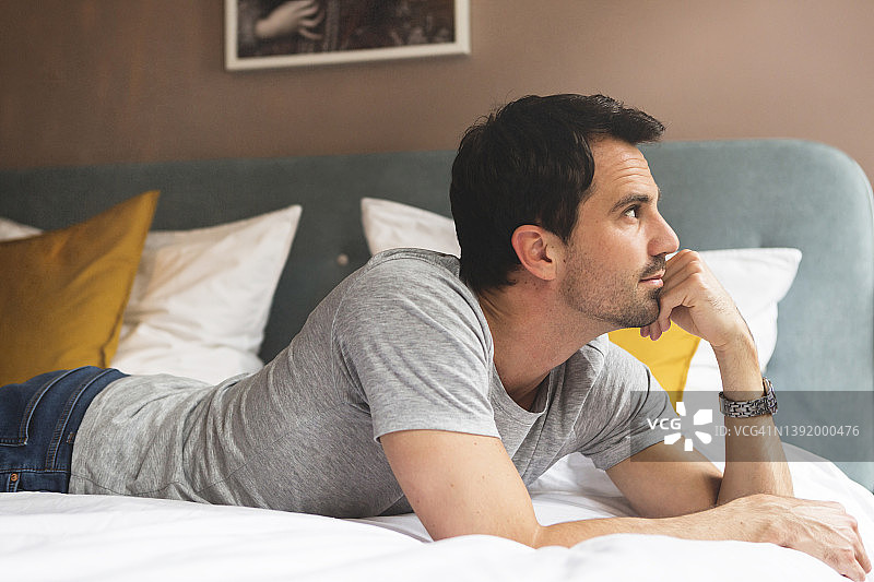 沉思的英俊男人躺在床上，在公寓里望着远处图片素材