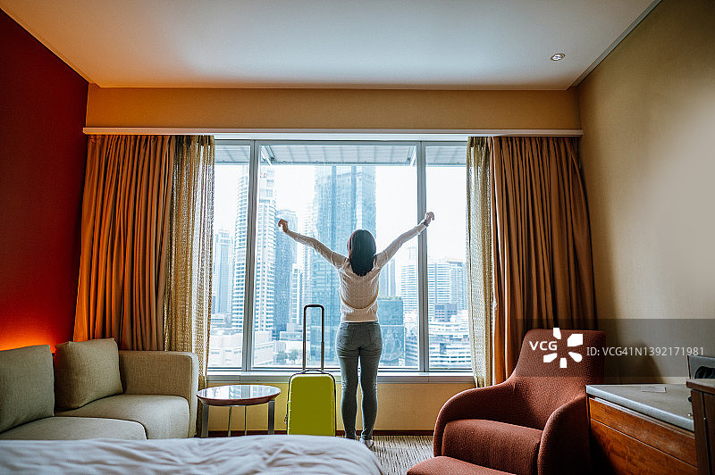 亚洲女游客办理入住后在酒店卧室，在窗边欣赏美景，举起双手，带着行李 - 单人旅行概念图片素材