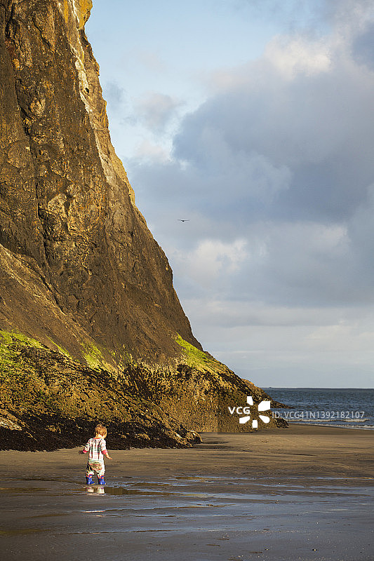 穿着靴子的小孩站在高耸悬崖下的海滩上图片素材