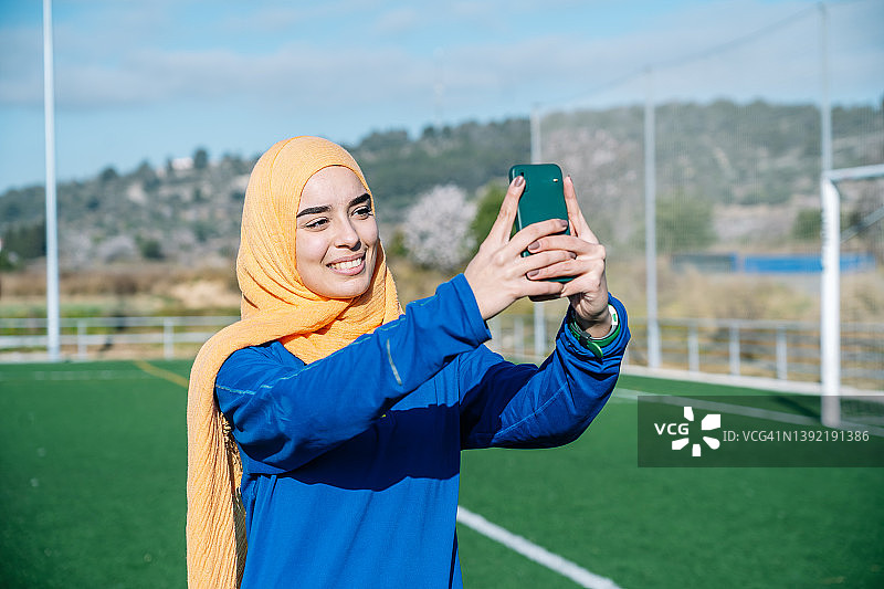 快乐的少数民族妇女在运动场上用智能手机自拍图片素材