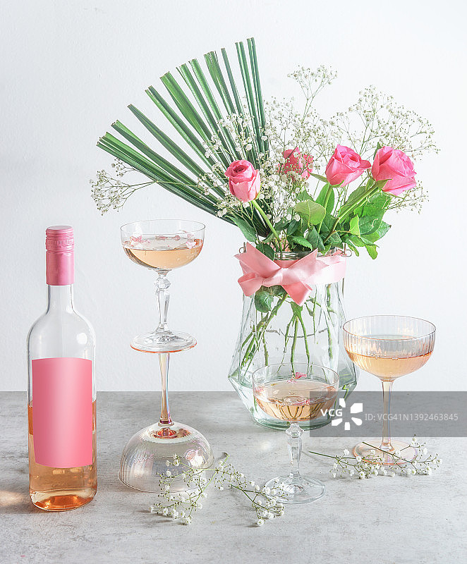 灰色厨房桌上的玫瑰葡萄酒瓶（带空白标签样机），香槟酒杯和白色墙壁背景上的花束图片素材