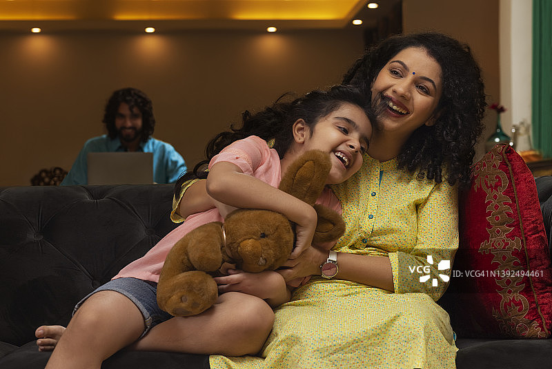 母亲在沙发上拥抱女儿，父亲在远处看着他们图片素材