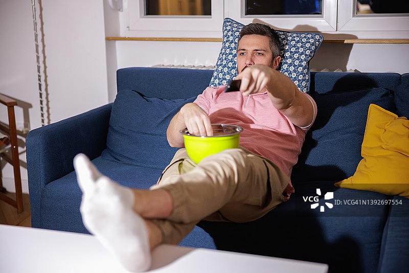 男人坐在家里的沙发上，一边吃零食一边用遥控器看电视图片素材