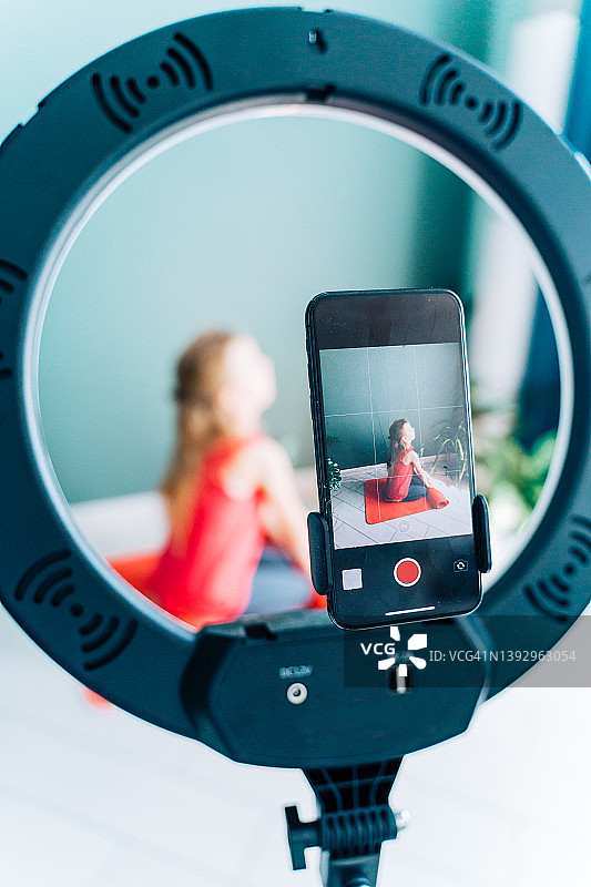 无线三脚架LED环形灯特写：拍摄金发小女孩上瑜伽课，坐在红色运动垫上图片素材