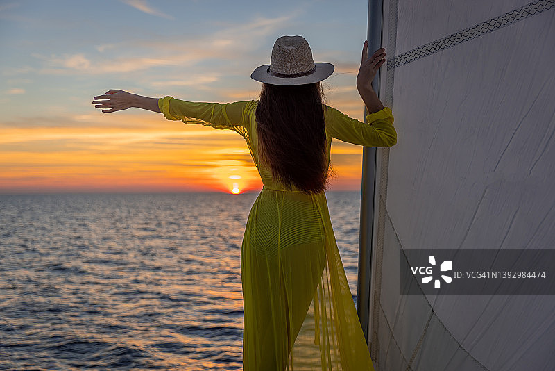 戴着帽子和穿着黄色连衣裙的长棕发女人站在帆船甲板上，看着日落图片素材