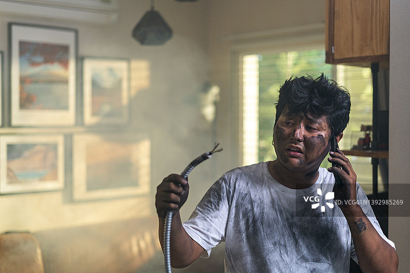 在家发生事故后，拿着电缆，脸脏兮兮、烧焦的滑稽表情，用手机绝望地打电话求助的年轻男子肖像。电力维修和DIY概念图片素材