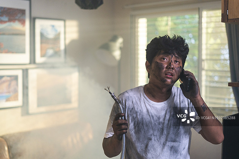 在家发生事故后，拿着电缆，脸脏兮兮、烧焦的滑稽表情，用手机绝望地打电话求助的年轻男子肖像。电力维修和DIY概念图片素材