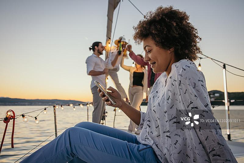 在帆船甲板上，一位棕色卷发的女士看着手机微笑，她的朋友们在背景中举行派对，举杯庆祝图片素材