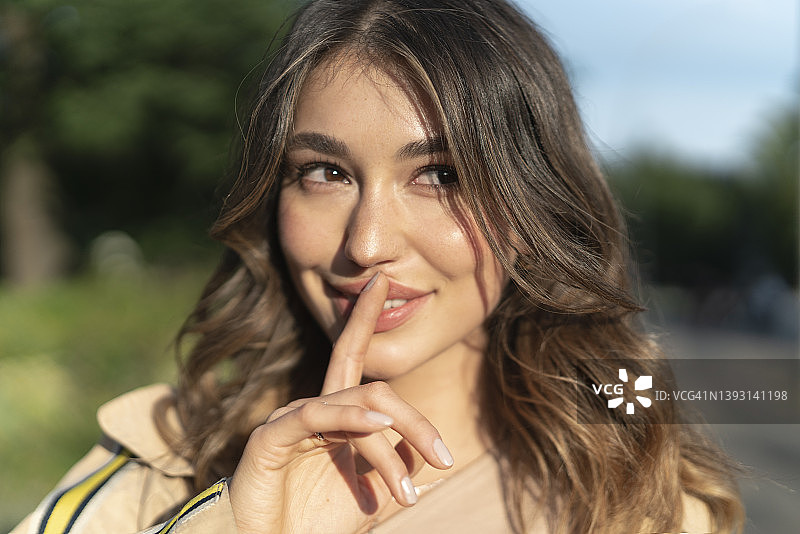 微笑的年轻棕发女子面部特写肖像，食指放在上唇图片素材