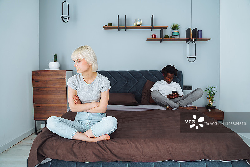 金发女人在和男友争吵后坐在床角。微笑的男人在上网，用智能手机与其他人聊天。手机成瘾图片素材