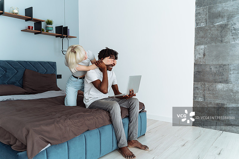 金发女人蒙住赤脚男友的眼睛，两人坐在床上使用银色笔记本电脑图片素材
