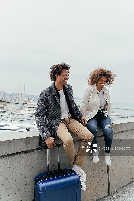 年轻的黑人夫妇笑着坐在墙上，旁边放着手提箱图片素材
