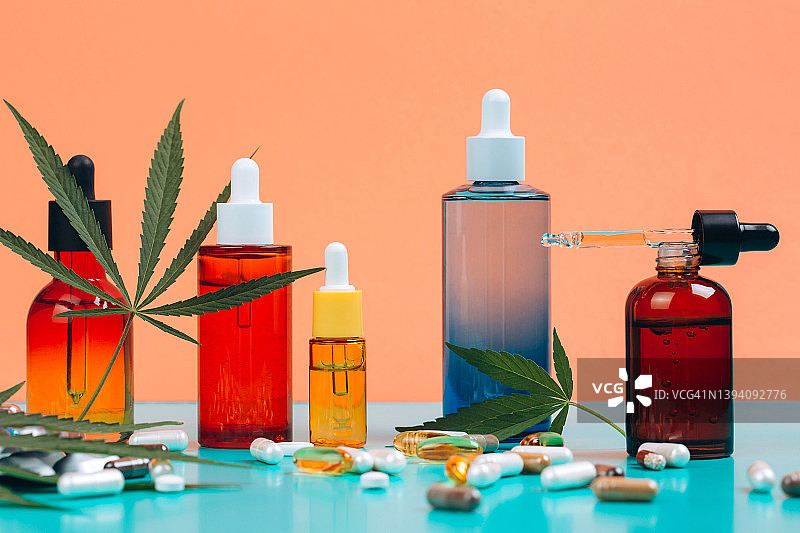 CBD油和营养补充剂套装，含大麻成分，橙色-蓝绿色背景，配大麻叶。五种彩色玻璃瓶一字排开，正面视角图片素材