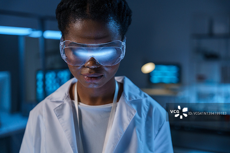 年轻漂亮的非洲裔美国女实验助理戴着防护眼镜向下看，在现代实验室里工作，特写图片素材