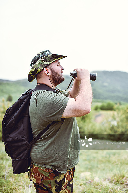 留着胡须的超重徒步者用双筒望远镜看风景的侧面照图片素材