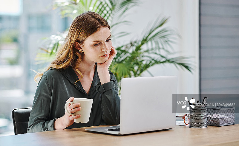 在办公室独自用笔记本电脑工作、喝咖啡和思考的年轻白人女商人图片素材