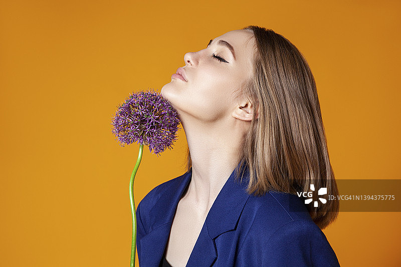 年轻女子闭眼用下巴触碰葱属植物，背景为橙色图片素材