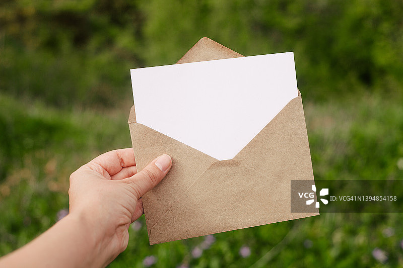 手中拿着带有复制空间的空白白纸卡的信封，背景是绿叶和鲜花图片素材
