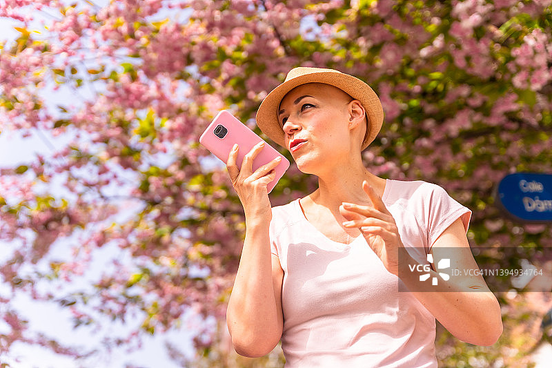 光头、戴着帽子、穿着粉色T恤的白人妇女在乳腺癌后享受生活。无发女孩在治愈疾病后微笑。发送语音消息图片素材