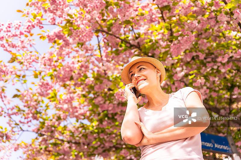 光头白人女性戴着帽子，身穿粉色T恤，乳腺癌幸存者享受生活，无发女孩治愈后微笑，正在打电话图片素材