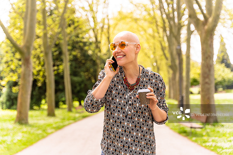 一位在乳腺癌后享受生活的秃顶白人妇女，戴着太阳镜在春天漫步公园，笑着打电话图片素材