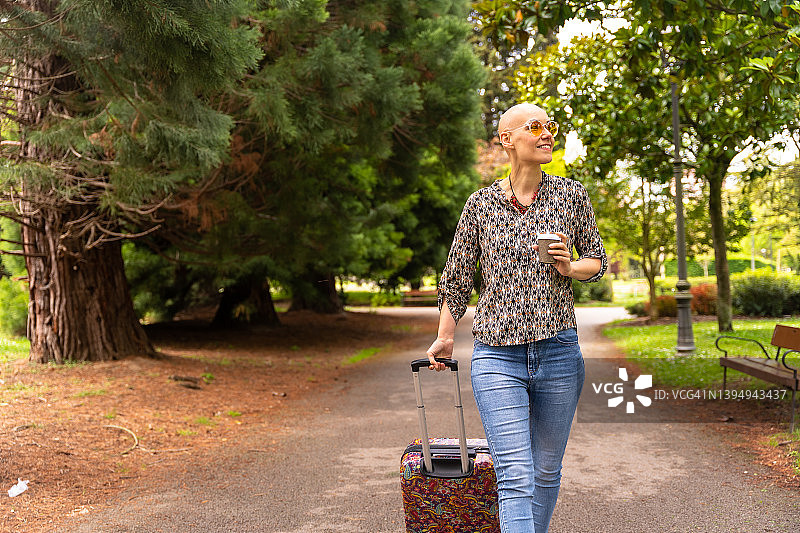 微笑的白人秃顶妇女带着行李箱和外卖咖啡在度假，享受乳腺癌后的生活，戴着太阳镜走过公园图片素材
