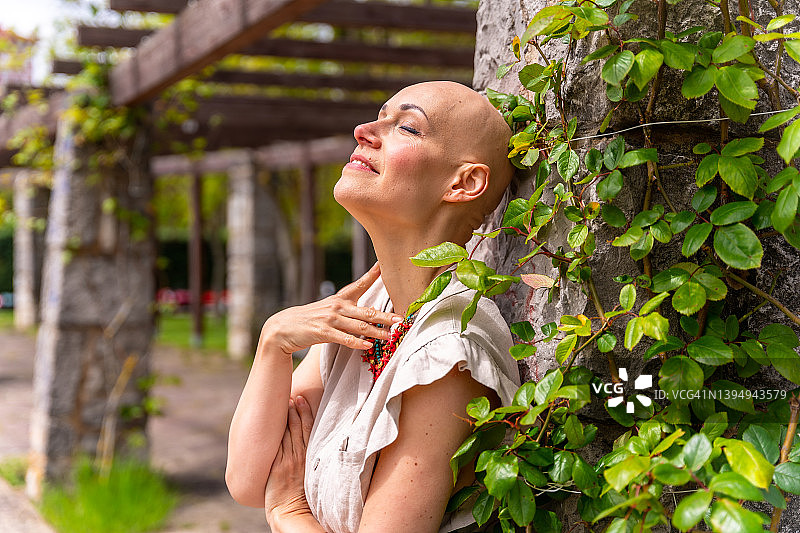 在春天公园里，一位患有脱发症的白人妇女在乳腺癌后享受生活，她闭着眼睛享受阳光图片素材
