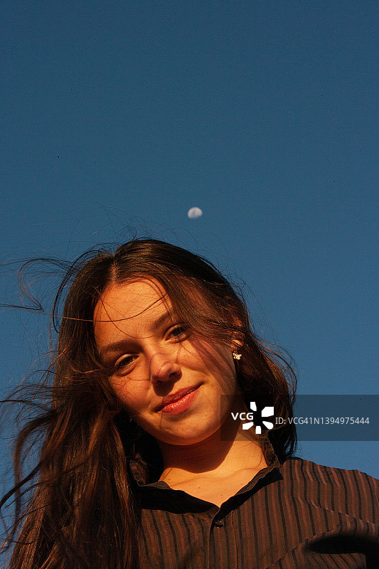一位年轻漂亮的女人，一个少女低头看着镜头，长长的深棕色头发在风中飘动，背景是晴朗的蓝天和皎洁的月亮图片素材