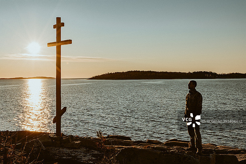 在阳光照耀的湖岸边，站在东正教十字架旁敬拜的男人剪影图片素材