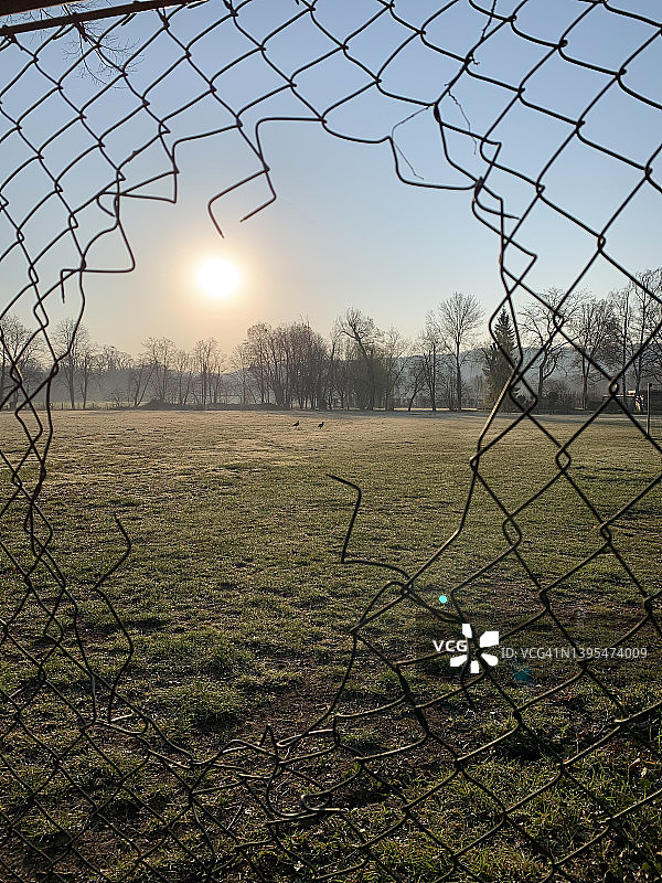 在春季一个寒冷的清晨，透过铁丝网链环围栏上的一个洞看到的运动场图片素材