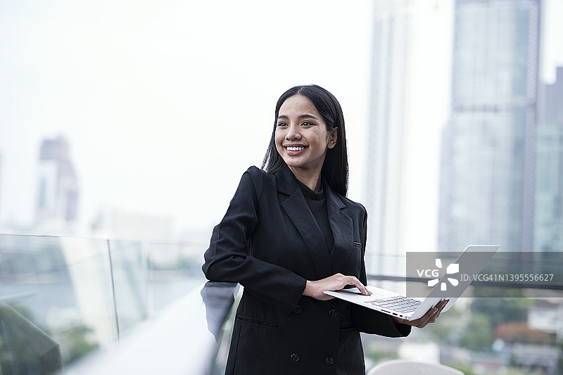 科技让沟通更轻松：自信的年轻女商人在酒店酒吧屋顶户外扶手上拿着笔记本电脑并看向远方图片素材