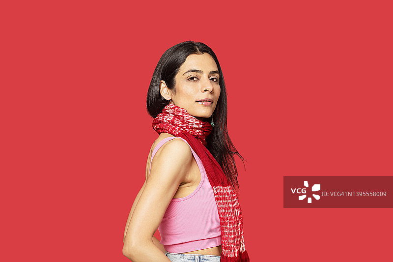 一位时尚的印度千禧一代女性，快乐地戴着红色围巾、粉色背心和绿松石耳环，站在工作室的红色背景前。图片素材