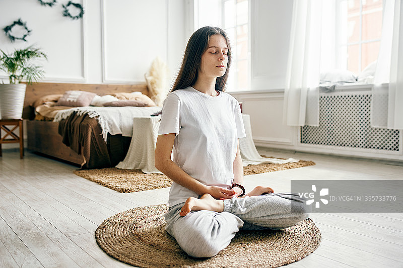 年轻漂亮的女人坐在明亮的房间里，在地毯上闭目冥想图片素材